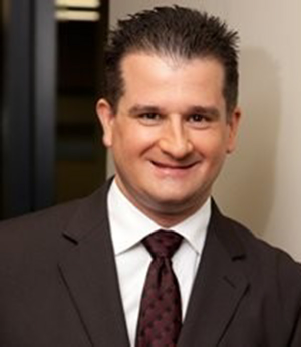 Bruce Adanac, a man with brown hair in a brown suit jacket and a brown tie smiles.